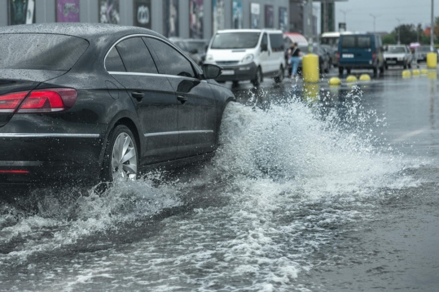 A black car drives through a large puddle on a wet city street, splashing water. Other vehicles and yellow bollards are visible in the background on the rainy day, capturing a scene familiar to any Denver engineer navigating urban rain.