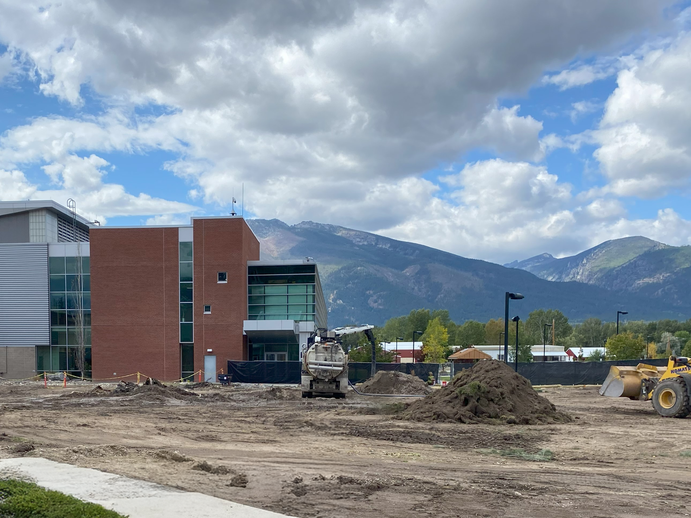 A construction site with dirt piles and machinery in front of a modern building, set against a backdrop of mountains under a partly cloudy sky.