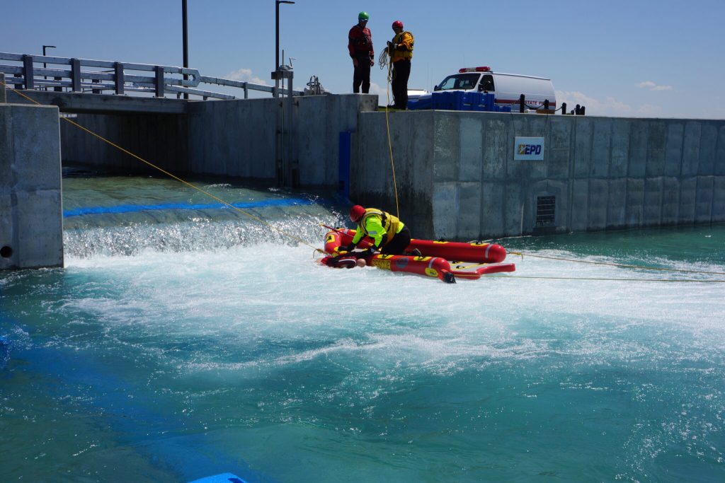 Two rescue workers in bright gear use an inflatable raft near a concrete water facility with fast-flowing water, while two others—possibly whitewater engineers—stand above on the platform with ropes, preparing for a water rescue operation.
