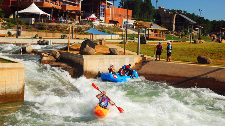 A kayaker paddles through whitewater rapids while a group in a blue raft navigates nearby. People watch from the grassy bank, and modern buildings—likely designed by a Denver engineer—rise in the background under a sunny sky.