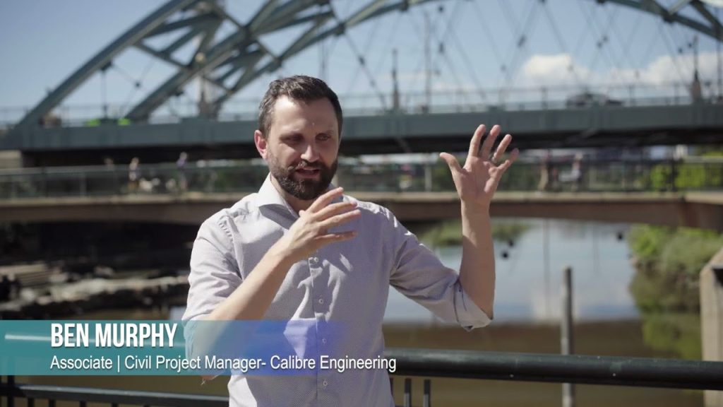 A man with a beard gestures while speaking outdoors near a river and a steel bridge. Text reads: Ben Murphy, Associate | Civil Project Manager - Calibre Engineering, a leader in Colorado engineering solutions.