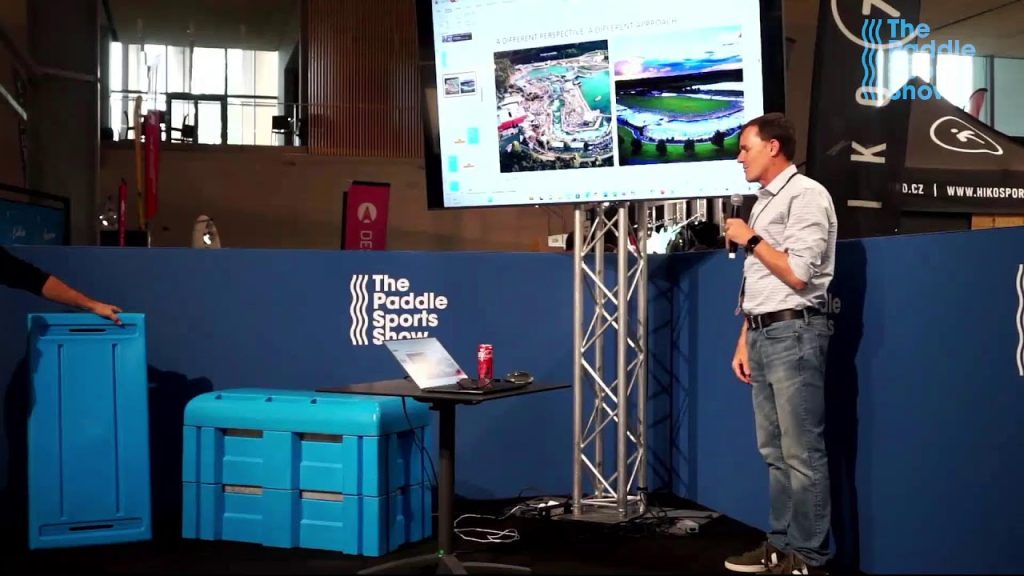 A man, possibly a Denver engineer, stands with a microphone, presenting slides at The Paddle Sports Show. Another person interacts with a blue container while a laptop and drink can sit on a table in front of the presenter.