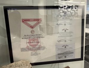 A glass display case contains a framed document with red and black text, a rough stone, a small architectural model, and other papers, all set in a modern, well-lit room with windows and reflections visible.