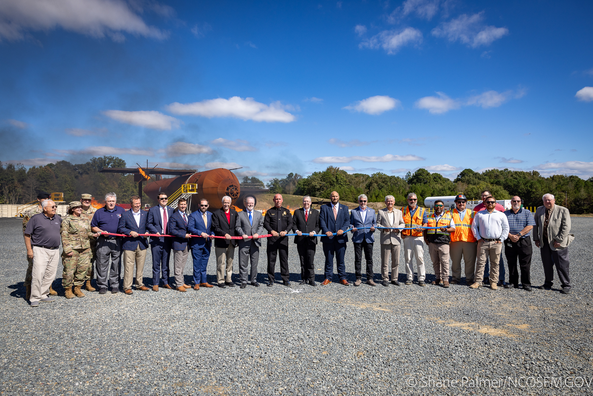 A group of people in business attire and uniforms stand in a line outdoors, holding a long red and blue ribbon at a ribbon-cutting ceremony on a gravel area, with construction equipment and trees in the background.