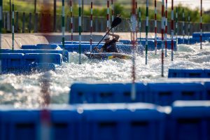 A kayaker navigates through whitewater rapids and slalom gates on an outdoor course, surrounded by blue barriers and splashing water under sunlight.