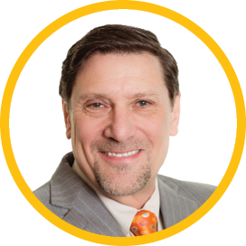 A smiling middle-aged man with short dark hair and a goatee, wearing a gray pinstripe suit, white shirt, and an orange polka-dot tie, framed by a yellow circle on a white background.