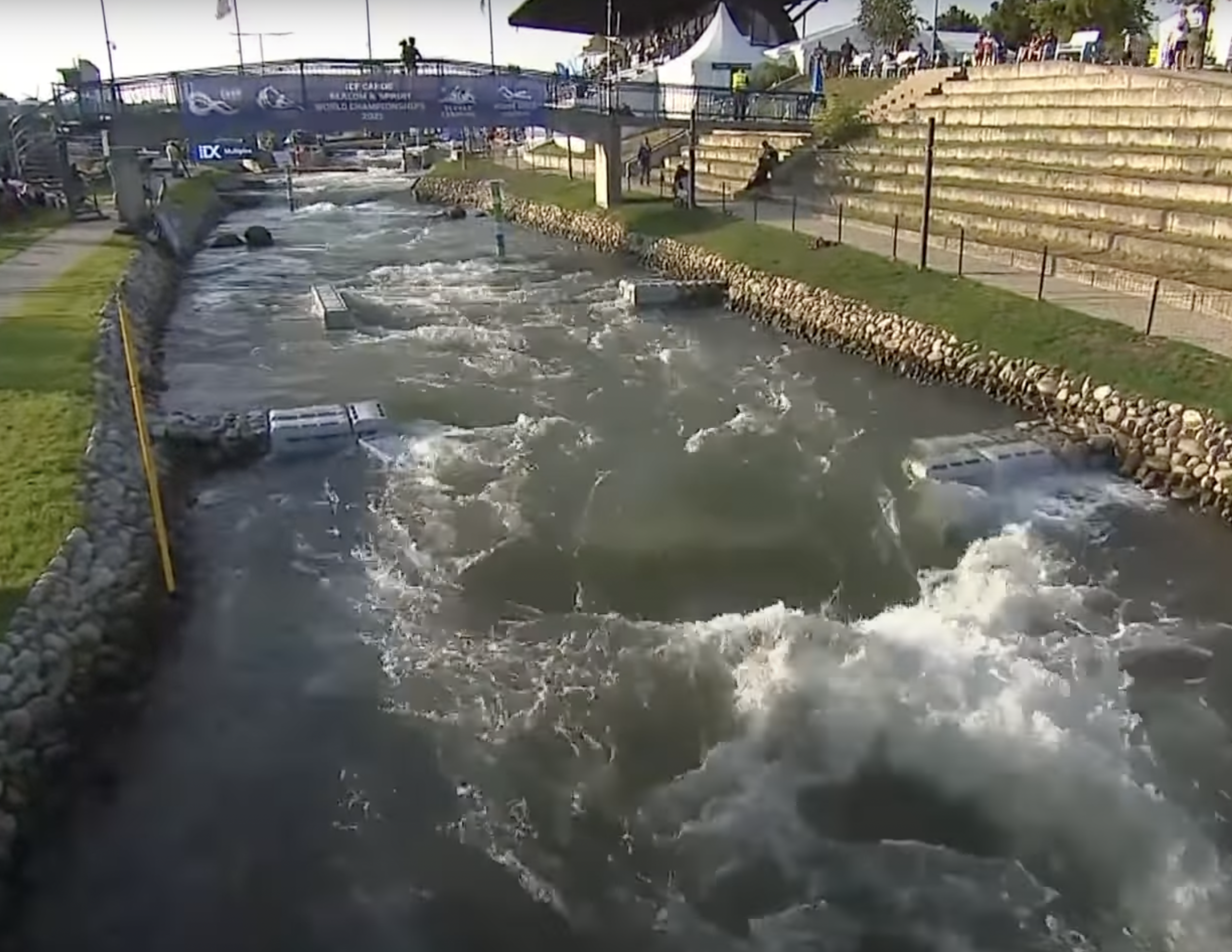 A man-made whitewater canoe slalom course with turbulent water, gates, stone banks, a grassy seating area, and a pedestrian bridge in the background under a blue banner.